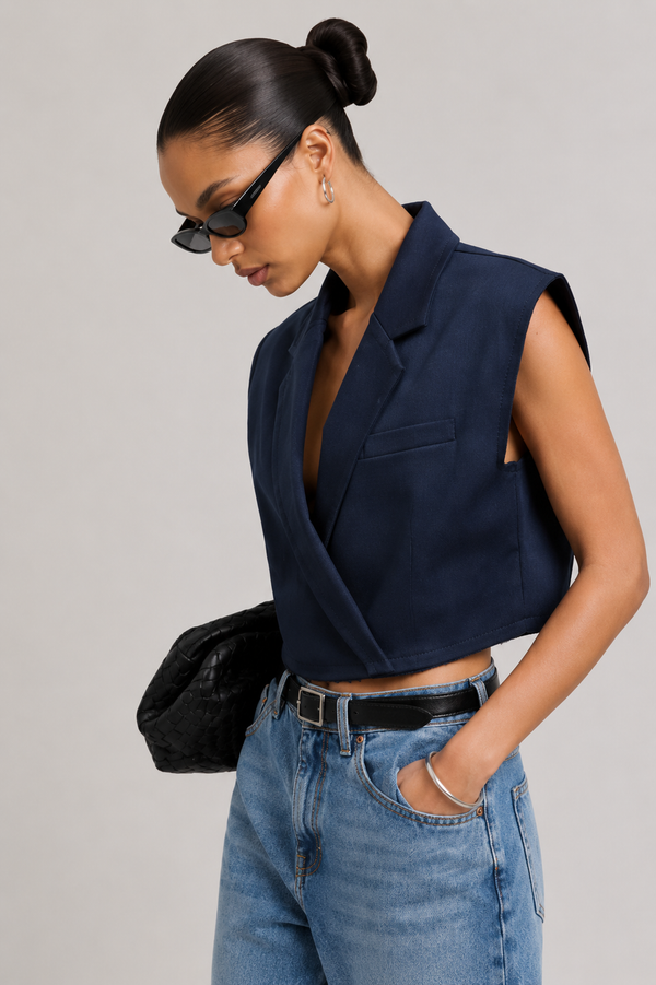 A woman wearing a navy blue sleeveless wrap-style blazer vest, light-wash blue jeans, a black belt, and black sunglasses, posed against a solid light gray background.