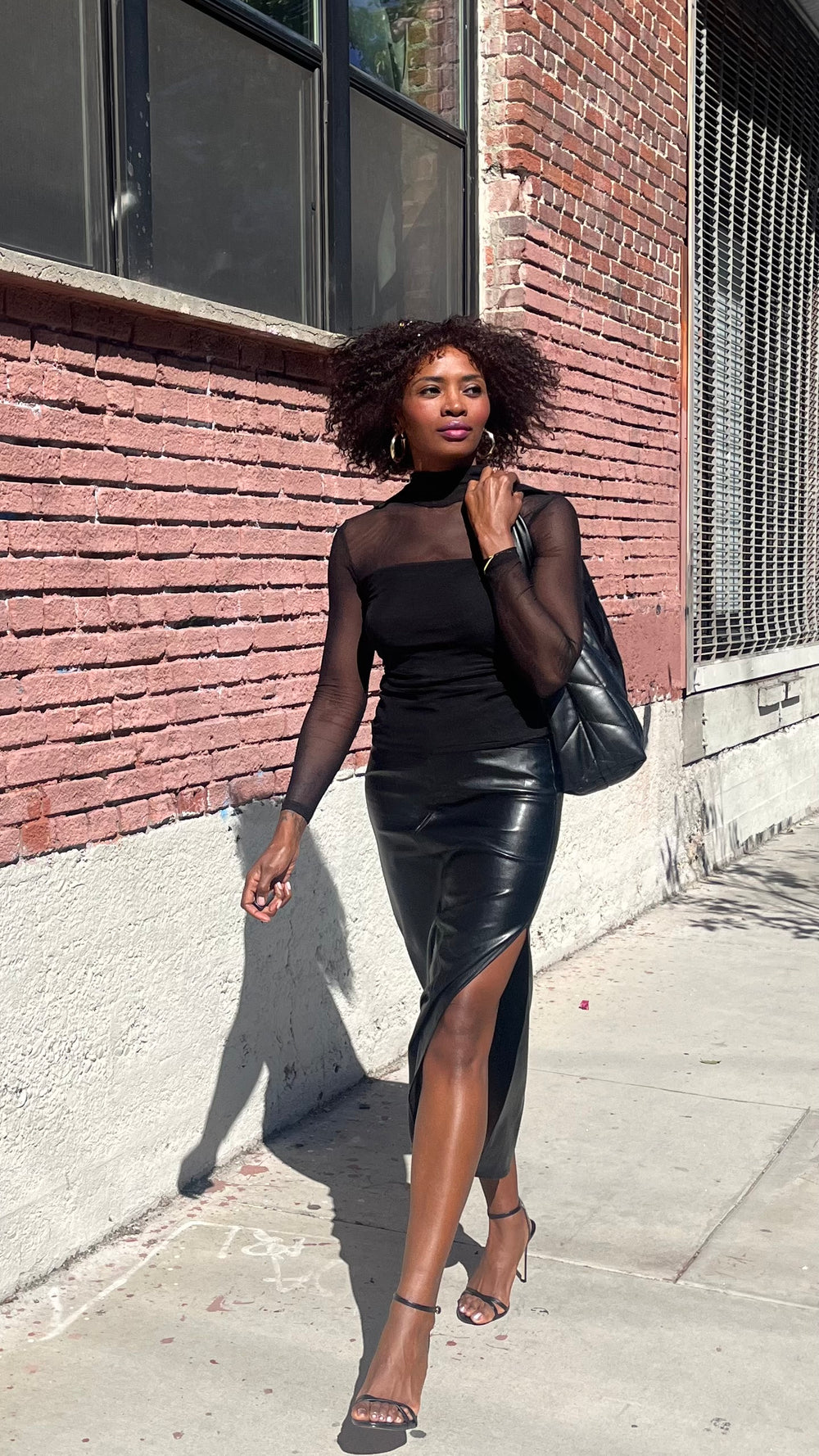 Woman in a black leather skirt and top standing against a brick wall.
