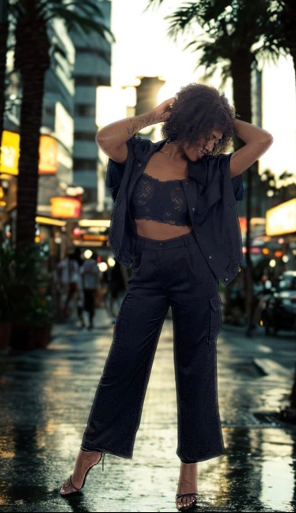 Woman in a stylish outfit walking on a city street with palm trees and lights in the background