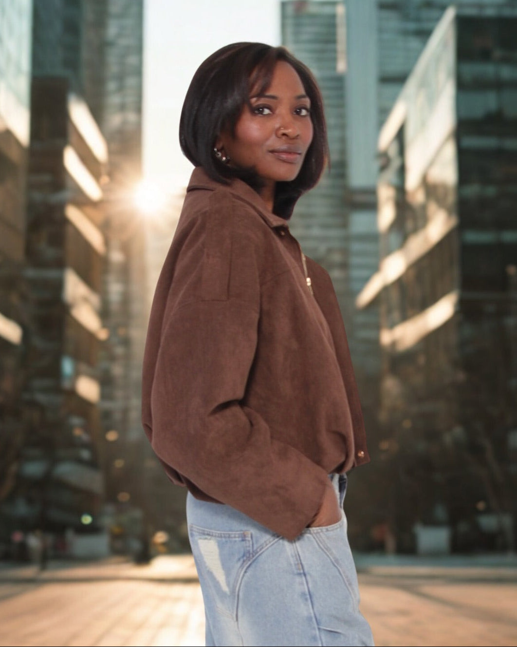 Woman in a brown jacket and light blue jeans standing in an urban setting with tall buildings.